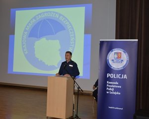 zastępca komendanta leżajskiej policji przemawia przy mównicy na tle wyświetlanego slajdu z napisem Krajowa Mapa Zagrożeń Bezpieczeństwa