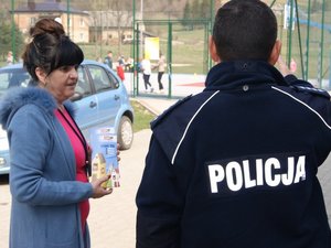policjant rozmawiający z kobietą która trzyma w ręku otrzymane materiały promocyjne, w tle bawiące się na boisku dzieci