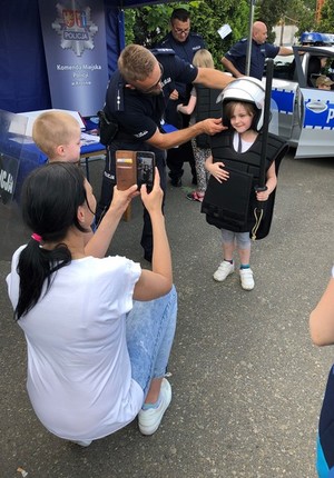 Na pierwszym planie kobieta która robi zdjęcie w tle policjant wraz z dziewczynką, który przymierza umundurowanie policyjne wykorzystywane do zabezpieczania imprez masowych.