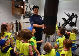 Fotografia kolorowa na której widać pomieszczenie siłowni. Na zdjęciu stoi policjantka a wokół niej dzieci ubrane w kamizelki odblaskowe.