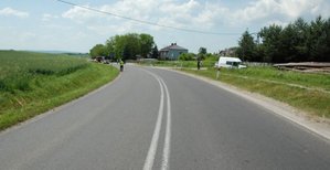 Fotografia kolorowa przedstawiająca miejsce wypadku drogowego. Na zdjęciu widoczna jest droga na której w oddali stoi umundurowany policjant, a za nim na drodze znajdują się pojazdy.