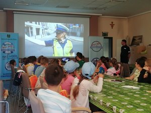Kolorowa fotografia. Dzieci siedzą przy stołach i oglądają film o Polskiej Policji. Film wyświetlany jest na ekranie. po obu stronach ekranu banery dotyczące akcji.