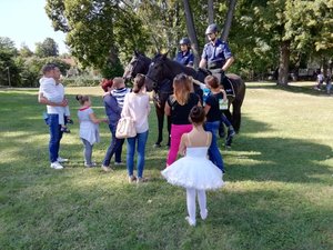 W środku funkcjonariusze na koniach policyjnych. Wokół stoją uczestnicy pikniku. Dzieci trzymane przez rodziców na rekach głaskają policyjne konie.