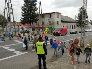Dzieci przechodzą przez przejście dla pieszych. Przed przejściem stoi policjant z założoną na mundur kamizelką odblaskową.