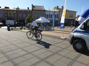 na fotografii funkcjonariusz w umundurowaniu policjanta z patrolu rowerowego, w tle radiowóz i rower policyjny na rynku starego miasta w Mielcu