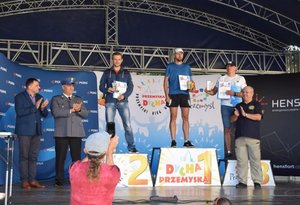 Na fotografii widoczna jest scena, na niej podium na którym znajdują się zwycięzcy biegu oraz osoby wręczające wyróżnienia i medale.