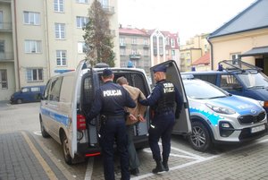 Na zdjęciu umundurowanie policjanci prowadza zatrzymanego do radiowozu celem przewiezienia do sądu na rozprawę.
