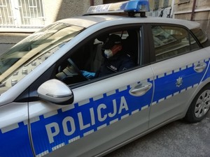 Policjant siedzący w radiowozie, ubrany w maseczkę ochronną oraz rękawiczki.