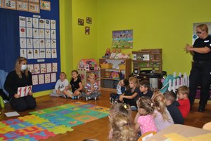 Zdjęcie kolorowe przedstawia klasę przedszkolną . Z prawej strony stoi policjantka apo lewej stronie pod ściana przednia jest druga policjantka. Dzieci siedzą w półkolu są ubrane w kolorowe rzeczy.Na podłodze rozłożone są kolorowe duże puzle