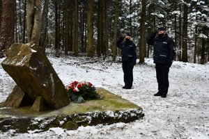 Złożenie kwiatów i zapalenie zniczy przy obelisku upamiętniającym katastrofę śmigłowca w Cisnej przez policjantów.  Policjant i policjantka (widok od przodu) oddają honory przed obeliskiem.