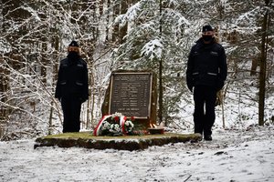 Złożenie kwiatów i zapalenie zniczy przy obelisku upamiętniającym katastrofę śmigłowca w Cisnej przez policjantów. Policjant i policjantka (widok od przodu) stoją obok obelisku.