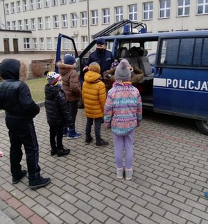 Policjant stojący przy radiowozie policyjnym. Na pierwszym planie szóstka dzieci odwrócona tyłem. W tle budynek szkoły.