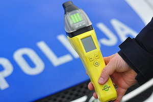 Zdjęcie kolorowe przedstawia przednia pokrywę silnika samochodu oznakowanego z białym napisem POLICJA , przed radiowozem widoczna ręka policjanta który trzyma urządzenie do badania stanu trzeźwości w kolorze żółtym