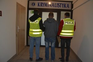 Na zdjęciu widoczne są sylwetki trzech mężczyzn, dwaj z nich mają zielone kamizelki z napisem policja. W tle przeszklone drzwi z napisem kryminalistyka