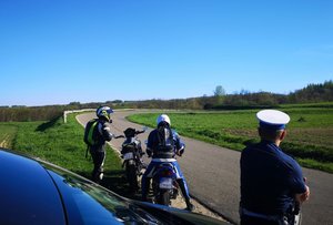 policjant w białej czapce oparty o samochód, przed nim, na poboczu dwaj motocykliści, jeden siedzi na motocyklu, drugi stoi