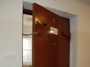 Kolorowa fotografia. Widok policyjnej celi w pomieszczeniu dla zatrzymanych. Otwarte drzwi metalowe do celi z założonym łańcuchem ograniczającym.