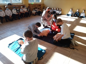 Na zdjęciu znajdują się uczniowie klękający przy fantomach leżących na materacach oraz ratowniczka medyczna na sali gimnastycznej