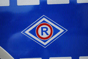 Oznaczenie służby ruchu drogowego, duża litera R wpisana w niebiesko-biały romb na granatowym tle.