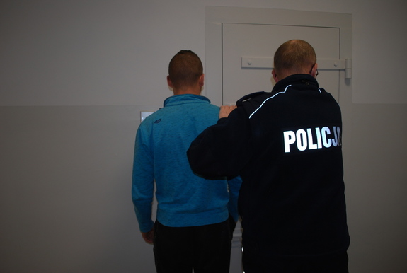Na zdjęciu w niebieskiej bluzie zatrzymany  mężczyzna, którego prowadzi umundurowany policjant. Obaj mężczyźni stoją odwróceni tyłem w pomieszczeniu la osób zatrzymanych.