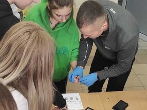 Na zdjęciu widoczna jest grupa młodzieży oraz technik kryminalistyki pobierający jednej z uczennic odcisk palca