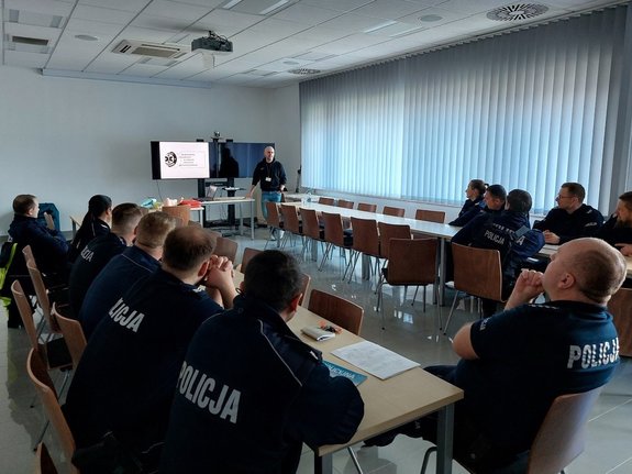 Uczestnicy szkolenia siedzą w sali konferencyjnej. Na ekranie monitora wyświetlany jest napis: „Doskonalenie umiejętności w zakresie udzielania pierwszej pomocy”. Obok stoi prowadzący szkolenie policjant.
