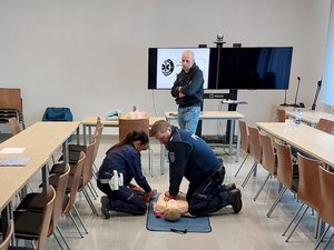 Policjant na leżącym na podłodze manekinie wykonuje masaż serca. Obok niego policjantka wypakowuje z przenośnej walizki automatyczny defibrylator zewnętrzny AED.