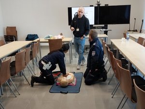 Policjantka na leżącym na podłodze manekinie wykonuje masaż serca. Jednocześnie słucha uwag przekazywanych jej przez prowadzącego szkolenie.