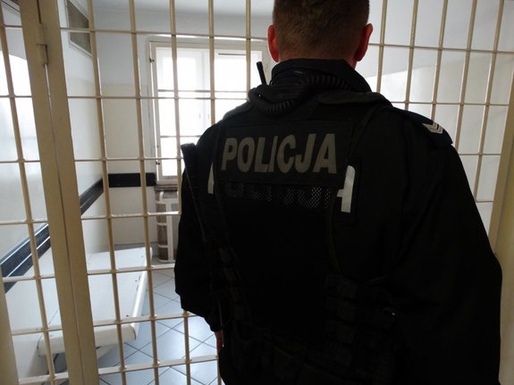 Na zdjęciu umundurowany policjant, stoi obok policyjnej izby zatrzymań