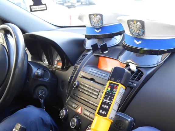 Wnętrze policyjnego radiowozu, na pulpicie, na podszybiu, leżą dwie policyjne czapki,  widoczne jest także urządzenie do badania stanu trzeźwości