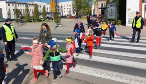 Funkcjonariusze Komendy Powiatowej Policji w Ropczycach asekurują podczas przejścia dzieci przez przejście dla pieszych. Na pasach znajduje się kilkoro dzieci w towarzystwie trójki opiekunów. Do przejścia do pieszej zbliża się kolejna grupka dzieci również w towarzystwie opiekunów.