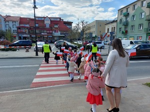 Na pierwszym planie widoczne jest przejście dla pieszych zlokalizowane przy ulicy Rynek w Ropczycach. Na przejściu znajduje się kilkanaście dzieci niosących chorągiewki w kolorze biało-czerwonym. Przed dziećmi jak również za nimi  idą  ich opiekunki. Z lewej i prawej strony dzieci idą funkcjonariusze policji. Po prawej stronie przed przejściem dla dzieci zatrzymał się kierujący pojazdu. W oddali widoczne budynki znajdujące się przy ulicy rynek w Ropczycach.