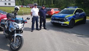 Policjanci - dzielnicowy i funkcjonariusz ruchu drogowego stoją obok oznakowanego radiowozu i motocykla
