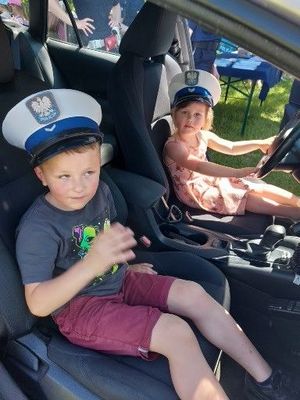 Na zdjęciu dzieci siedzące w radiowozie, na głowach maja założone białe policyjne czapki.