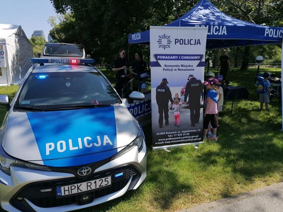 Na zdjęciu policyjny radiowóz, obok baner Komendy Miejskiej Policji w Rzeszowie  oraz namiot z napisem policja.