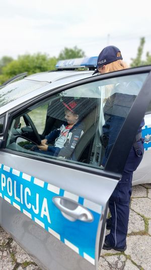 Chłopiec siedzi w radiowozie na miejscu kierowcy, policjantka stoi obok samochodu.