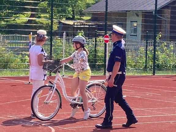 Uczestniczka egzaminu na kartę rowerową stoi przed linią z napisem STOP. Obok niej stoi policjant oraz nauczycielka.