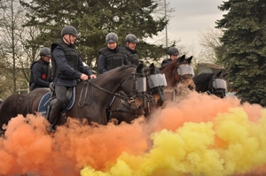 Umundurowani policjanci na koniach podczas atestacji na placu. Policjanci na koniach. Przed nimi kłęby dymu w kolorach pomarańczowym i żółtym.